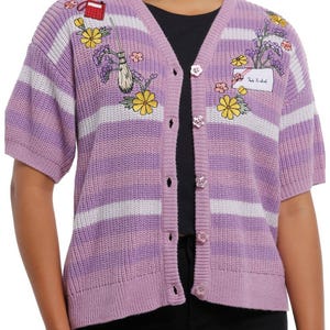 May include: A lilac short-sleeve cardigan with white stripes and floral embroidery. The button-up sweater features embroidered flowers, a broom, and a small red bag. The text "Tale Untold" is on a patch.