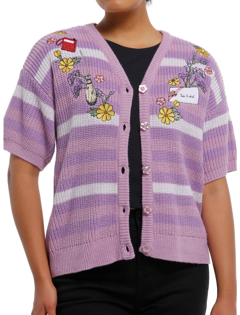 May include: A lilac short-sleeve cardigan with white stripes and floral embroidery. The button-up sweater features embroidered flowers, a broom, and a small red bag. The text "Tale Untold" is on a patch.