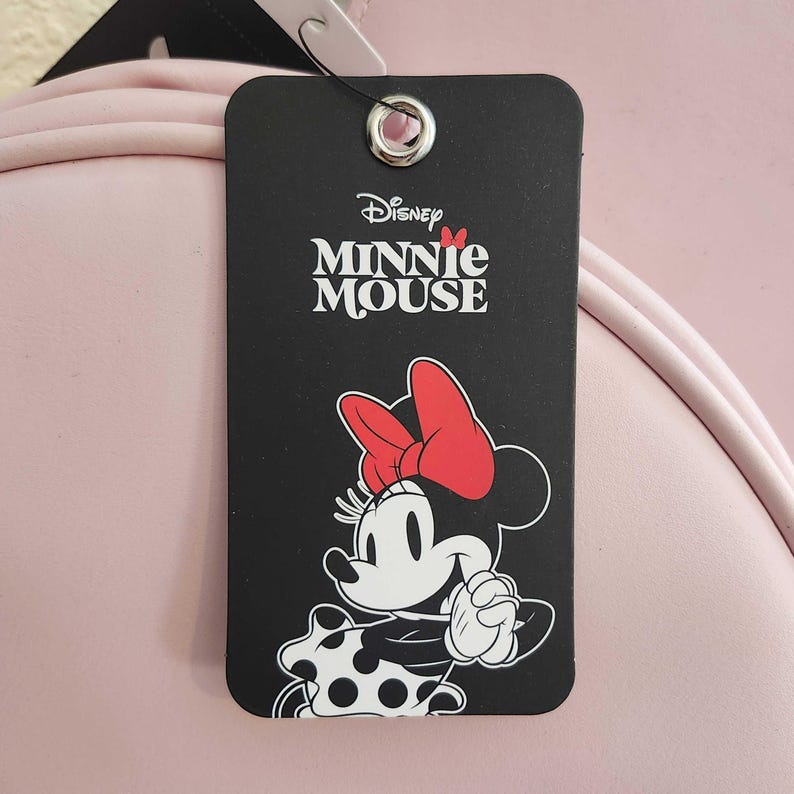 May include: A black tag featuring the Disney Minnie Mouse logo and a cartoon illustration of Minnie Mouse. Minnie Mouse is depicted with a red bow, a polka-dot skirt, and white gloves. The tag is attached to a pink object.