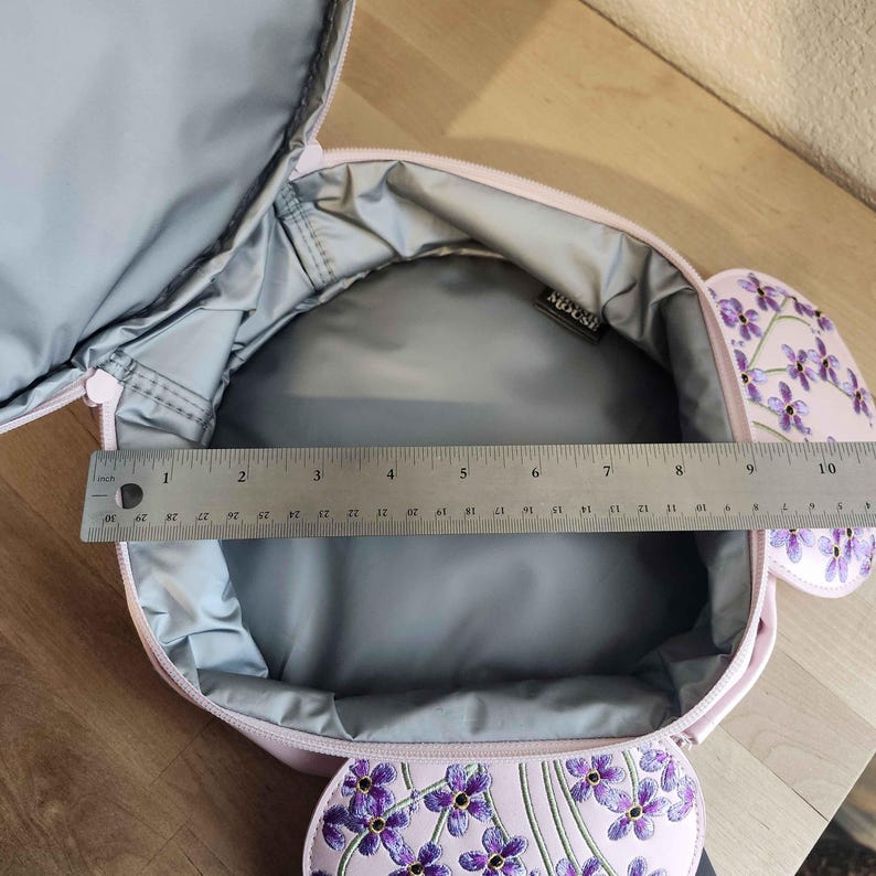 May include: Open pink lunch bag with a floral design featuring purple flowers. The interior is gray, and a ruler shows the bag's width is approximately 10 inches. The bag has a zipper closure and a tag that says "MOUSE".