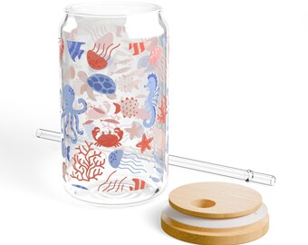 May include: A clear glass can-shaped drinking glass with a sea life pattern, including red crabs, blue octopuses, and seahorses. A clear glass straw and a wooden lid with a white insert are included. The glass is designed for cold beverages.