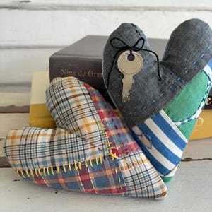 May include: Two stuffed fabric hearts. One heart features a multi-colored plaid pattern with yellow decorative stitching. The other heart is made of grey, green, and blue striped fabric with a silver metal key tied to it with black string.
