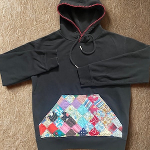 May include: Dark gray hoodie with a patchwork pocket featuring a variety of colorful fabric squares. The hood has red stitching around the edge, and a drawstring. A casual, comfortable garment.