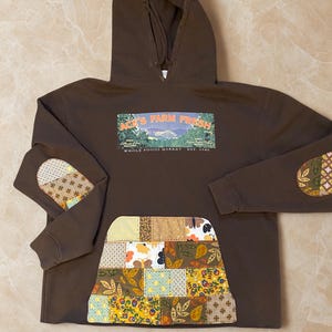 May include: Brown hoodie with a front pocket and elbow patches made of patchwork fabric. The hoodie features the text "Ace's Farm Fresh" and "Whole Foods Market Est. 1980" in a rectangular design on the chest.