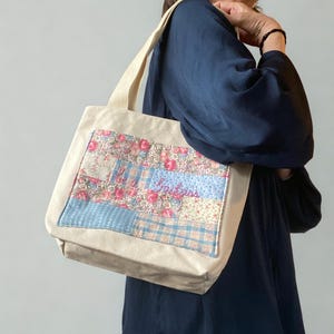May include: Beige tote bag with a floral patchwork design. The patchwork includes pink roses, blue plaid, and a blue stripe with embroidered text. The bag has beige canvas handles and a rectangular shape.