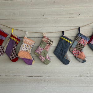 May include: A row of seven hanging Christmas stockings made from various fabric scraps, including denim, floral prints, gingham, and solid colors. Each stocking features unique patchwork designs, decorative trim, and hanging loops attached to a twine string.