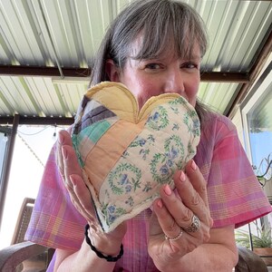 May include: A heart-shaped fabric ornament constructed from quilted patchwork panels. The design features a mix of light blue, peach, and cream-colored fabric strips alongside a section of white fabric with a blue floral wreath pattern.