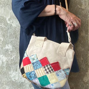 May include: Cream-colored tote bag with a patchwork design. The patchwork features squares of red, blue, and patterned fabrics. The bag has two short handles and a long, adjustable strap. The bag is held by a person.