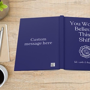 May include: A navy blue journal with the text "You Won't Believe This Shift" and a fire department emblem. The left side of the journal reads "Custom message here". A spiral notebook, pen, and coffee cup are also on the wooden surface.