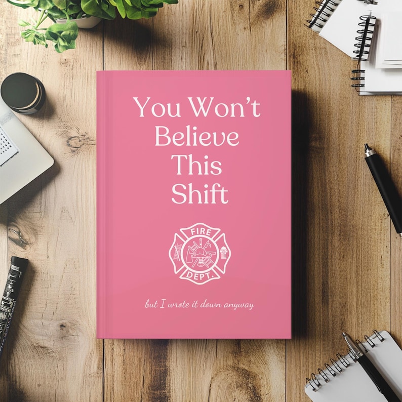 May include: A pink notebook with the text "You Won't Believe This Shift" and a fire department emblem. The notebook also has the text "but I wrote it down anyway". The notebook is on a wooden surface.