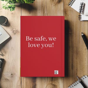 May include: A red journal with the text "Be safe, we love you!" in white. The journal is on a wooden surface, surrounded by office supplies and a small plant.