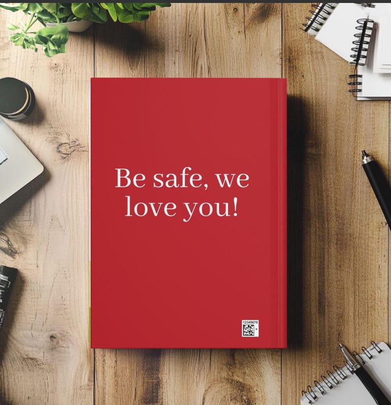 May include: A red journal with the text "Be safe, we love you!" in white. The journal is on a wooden surface, surrounded by office supplies and a small plant.