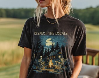 Może przedstawiać: Czarny t-shirt z krótkim rękawem z grafiką przedstawiającą dwa szkielety siedzące przy ognisku, niedźwiedzia i chatę w lesie pod pełnią księżyca. Napis "RESPECT THE LOCALS" jest nadrukowany w jasnobeżowym kolorze nad ilustracją graficzną.