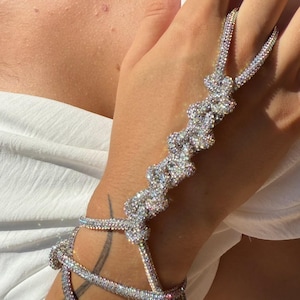 May include: A sparkling silver hand chain bracelet. The bracelet features multiple strands of shimmering rhinestones that connect to a finger ring and a wrist cuff. The jewelry is worn on a hand with red nail polish and a white top.
