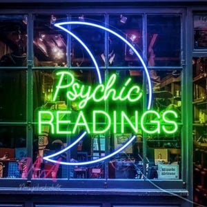 May include: A neon sign in a window displays the words "Psychic READINGS" in bright green and white. The sign is framed by a blue crescent moon shape. The window reveals a glimpse of a shop interior.
