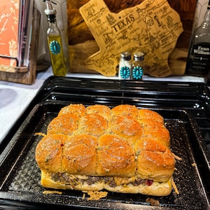 Puede incluir: Una bandeja de sliders horneados con tapas doradas, rellenos de carne y queso. Los sliders est&aacute;n en una bandeja para hornear negra. En el fondo, se ve una tabla de cortar de madera con forma de Texas y una botella de aceite de oliva.