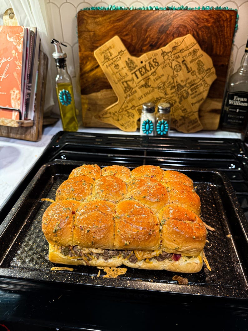 Puede incluir: Una bandeja de sliders horneados con tapas doradas, rellenos de carne y queso. Los sliders est&aacute;n en una bandeja para hornear negra. En el fondo, se ve una tabla de cortar de madera con forma de Texas y una botella de aceite de oliva.