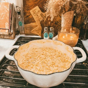 Puede incluir: Una fuente blanca con bordes festoneados llena de un plato cremoso de ma&iacute;z de color dorado. El plato tiene dos asas y est&aacute; sobre una encimera negra. El fondo incluye un libro de cocina, tablas de cortar de madera y objetos decorativos.