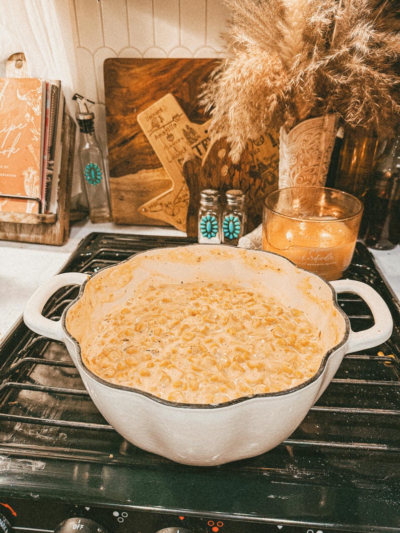 Puede incluir: Una fuente blanca con bordes festoneados llena de un plato cremoso de ma&iacute;z de color dorado. El plato tiene dos asas y est&aacute; sobre una encimera negra. El fondo incluye un libro de cocina, tablas de cortar de madera y objetos decorativos.