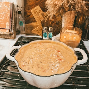 Puede incluir: Una fuente blanca con bordes festoneados llena de sopa cremosa, colocada sobre una estufa negra. La sopa tiene un color naranja claro con ingredientes visibles. El plato tiene dos asas. Otros elementos en el fondo incluyen un libro de cocina y objetos decorativos.