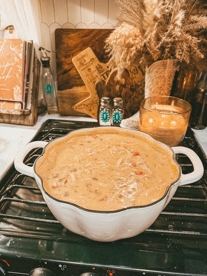 Puede incluir: Una fuente blanca con bordes festoneados llena de sopa cremosa, colocada sobre una estufa negra. La sopa tiene un color naranja claro con ingredientes visibles. El plato tiene dos asas. Otros elementos en el fondo incluyen un libro de cocina y objetos decorativos.