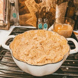 Puede incluir: Una fuente blanca con bordes festoneados llena de un plato cremoso beige se encuentra sobre una rejilla de horno negra. El plato tiene dos asas. Otros elementos en el fondo incluyen una tabla de cortar de madera, una vela y objetos decorativos.