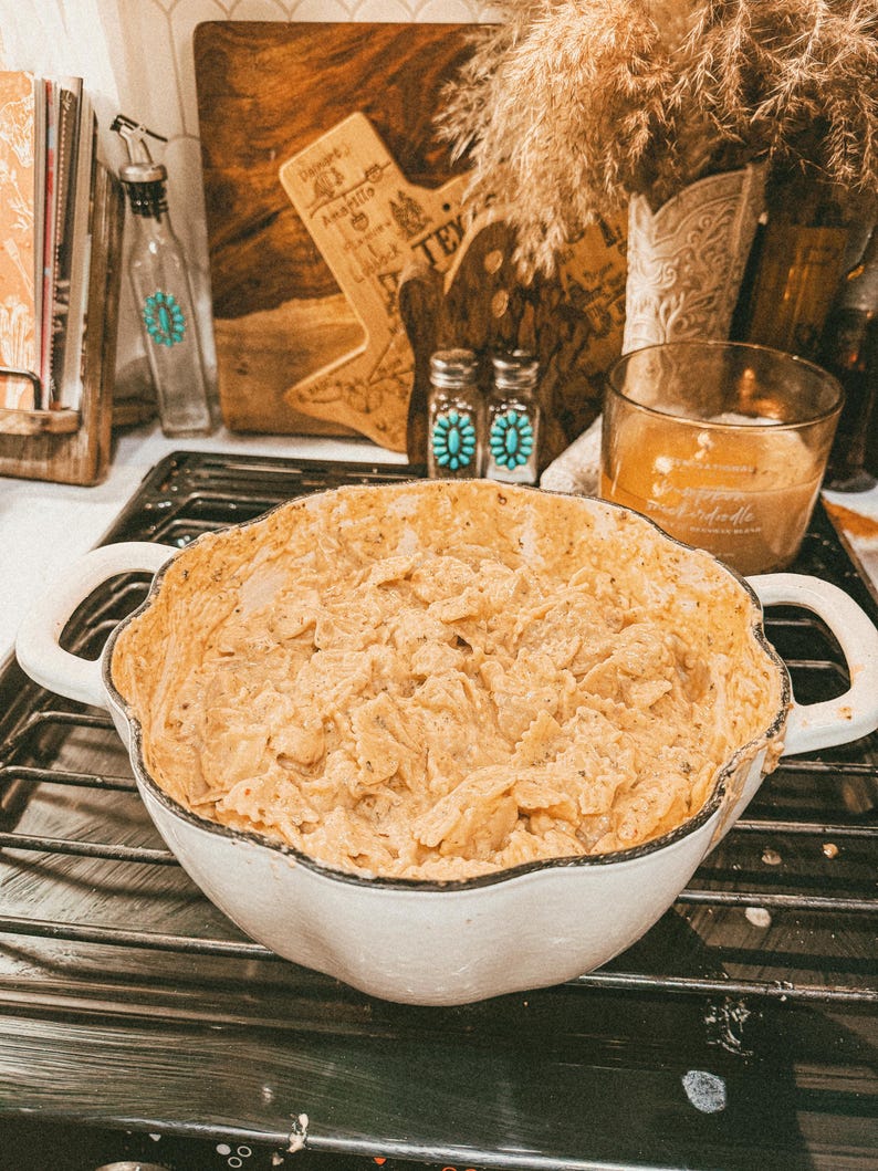 Puede incluir: Una fuente blanca con bordes festoneados llena de un plato cremoso beige se encuentra sobre una rejilla de horno negra. El plato tiene dos asas. Otros elementos en el fondo incluyen una tabla de cortar de madera, una vela y objetos decorativos.