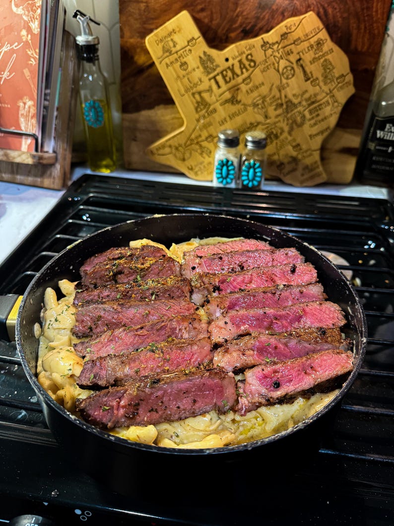 Puede incluir: Una sart&eacute;n con filete en rodajas cocinado con una salsa cremosa. El filete est&aacute; sazonado y tiene un interior rosado. Una tabla de cortar de madera con forma de Texas y saleros y pimenteros est&aacute;n en el fondo.