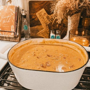 Puede incluir: Una olla de hierro fundido esmaltada blanca, llena de un guiso cremoso de color naranja, se encuentra sobre una estufa negra. La olla tiene dos asas blancas. En el fondo, un libro de cocina, una tabla de cortar de madera y objetos decorativos completan la escena.