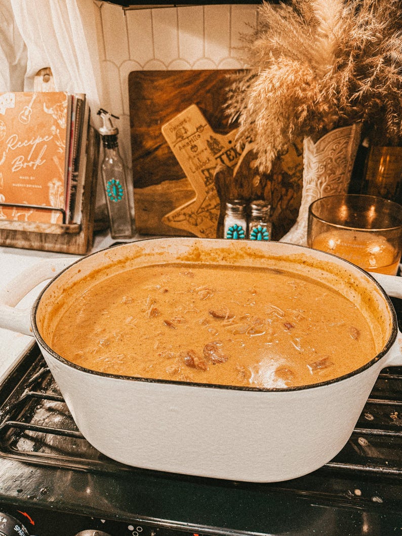 Puede incluir: Una olla de hierro fundido esmaltada blanca, llena de un guiso cremoso de color naranja, se encuentra sobre una estufa negra. La olla tiene dos asas blancas. En el fondo, un libro de cocina, una tabla de cortar de madera y objetos decorativos completan la escena.