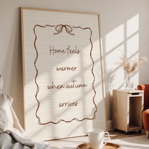 May include: A framed print with the text "Home feels warmer when autumn arrives" in brown script. The artwork has a cream background with a dotted pattern and a brown border. A white mug sits on a wooden tray in front of the print.