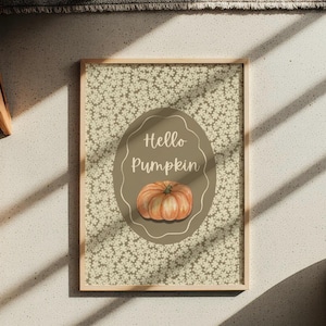 Può includere: Una stampa incorniciata con uno sfondo a motivi floreali e un design ovale centrale. L'ovale contiene il testo "Hello Pumpkin" sopra un'illustrazione ad acquerello di una zucca arancione. La cornice è color legno chiaro.