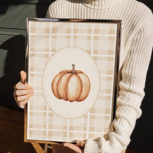 May include: A framed print with a pumpkin illustration. The pumpkin is orange, set within an oval frame. The background has a plaid pattern in beige and tan, creating an autumnal aesthetic. The frame has a metallic finish, adding a touch of elegance.