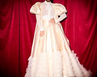Puede incluir: Una persona vistiendo un vestido blanco con volantes, con una parte superior transparente y una corbata de lazo blanca. El vestido tiene una falda completa y está hecho de tul. La persona está de pie frente a una cortina de terciopelo rojo.