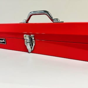 May include: A bright red metal toolbox with a silver handle and latch. The toolbox has the brand name "Howak" in a white rectangle on the side. The toolbox is rectangular and appears to be empty.