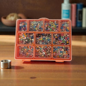 May include: An orange plastic organizer filled with various colorful beads, buttons, and small craft supplies. The organizer has multiple compartments, each holding different types of embellishments. A small silver metal ring sits on the wooden surface.