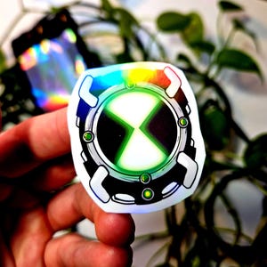 May include: A holographic sticker featuring a circular design with a black and white outer ring, a green and black center, and a white X-shaped symbol. The sticker has a rainbow-colored border and is surrounded by green foliage.