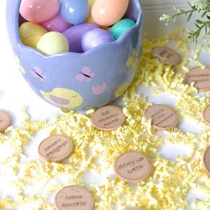 May include: A blue ceramic Easter basket filled with pastel plastic eggs. The basket is decorated with butterflies and a yellow chick. Scattered around the basket are small wooden circles with various phrases written on them, such as "Ice Cream Date", "Craft Project", and "Stay Up Late".