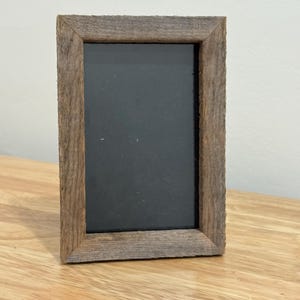 May include: A rectangular wooden picture frame with a dark grey interior. The frame is made of weathered, light brown wood, and sits on a light brown wooden surface. The frame is empty.