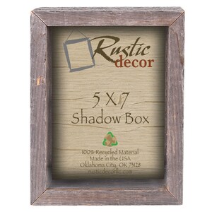 May include: A rustic wood shadow box frame with a 5x7 inch opening. The frame is made from 100% recycled material and is made in the USA. The frame is brown with a light wood grain and has a distressed look. The text "Rustic decor" is printed on the frame in red and brown lettering. The text "5x7 Shadow Box" is printed on the frame in brown lettering. The text "100% Recycled Material Made in the USA Oklahoma City, OK 73128 rustiedecorllc.com" is printed on the frame in black lettering.