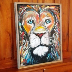 May include: A framed painting of a lion's face. The artwork features a colorful, expressive style with a focus on the lion's blue eyes, red nose, and white muzzle. The mane is depicted with vibrant brushstrokes of orange, yellow, and blue.