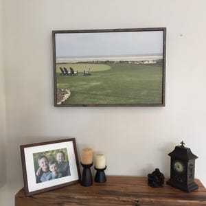 May include: A framed landscape print of a golf course with chairs overlooking the ocean. Below, a framed photo of three children, two candles, a black clock, and a decorative object sit on a wooden table.