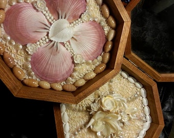 Könnte beinhalten: Eine sechseckige Holzkiste mit zwei Fächern, die jeweils mit einem Mosaik aus Muscheln verziert sind. Das obere Fach zeigt ein Design aus rosa und weißen Muscheln, während das untere Fach ein Design aus weißen Muscheln zeigt.