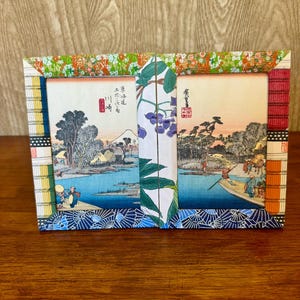 May include: A two-part picture frame made of colorful patterned paper. The frame is decorated with a floral design and has a blue and white patterned border. The frame is holding two Japanese prints of landscapes.