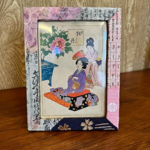 May include: A framed Japanese print featuring a seated woman in a purple kimono, attended by another woman. The frame is decorated with patterned fabric and paper in shades of pink, purple, and blue. The print depicts a traditional scene with floral and script elements.