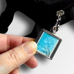 May include: Black small bag with a zipper, a teal camera, and a passport. A hand holds a small square charm with a blue stamp design. Text reads: Use as a keyring, zipper pull or handbag charm.