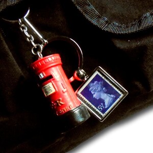 May include: A red miniature pillar box keychain with a silver chain and a small square charm featuring a blue postage stamp. The keychain is hanging against a dark background. The text on the image says "Your keychain will come with both split ring & lanyard clip (pillar box charm NOT included)".