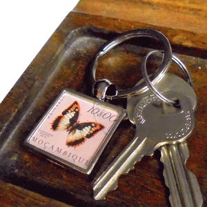 May include: A wooden key holder with a butterfly keychain and two keys. The keychain features a pink background with a butterfly image and the word "MOZAMBIQUE". The key holder has a dark brown finish.