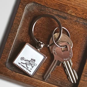 May include: A wooden key and coin tidy with a rectangular shape and a natural wood finish. The organiser has compartments for keys, coins, and a keychain with a graphic. The keychain has the text "US POSTAL SERVICE".