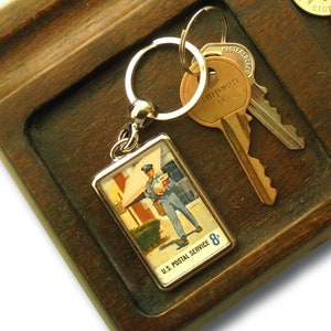 Könnte beinhalten: Schlüssel- und Geldtablett aus Holz mit zwei Fächern. Das Tablett hat eine dunkelbraune Beize und ist aus Holz. Ein Schlüsselanhänger mit einem kleinen Bild eines Briefträgers hängt am Tablett. Es befinden sich mehrere Münzen im Tablett.